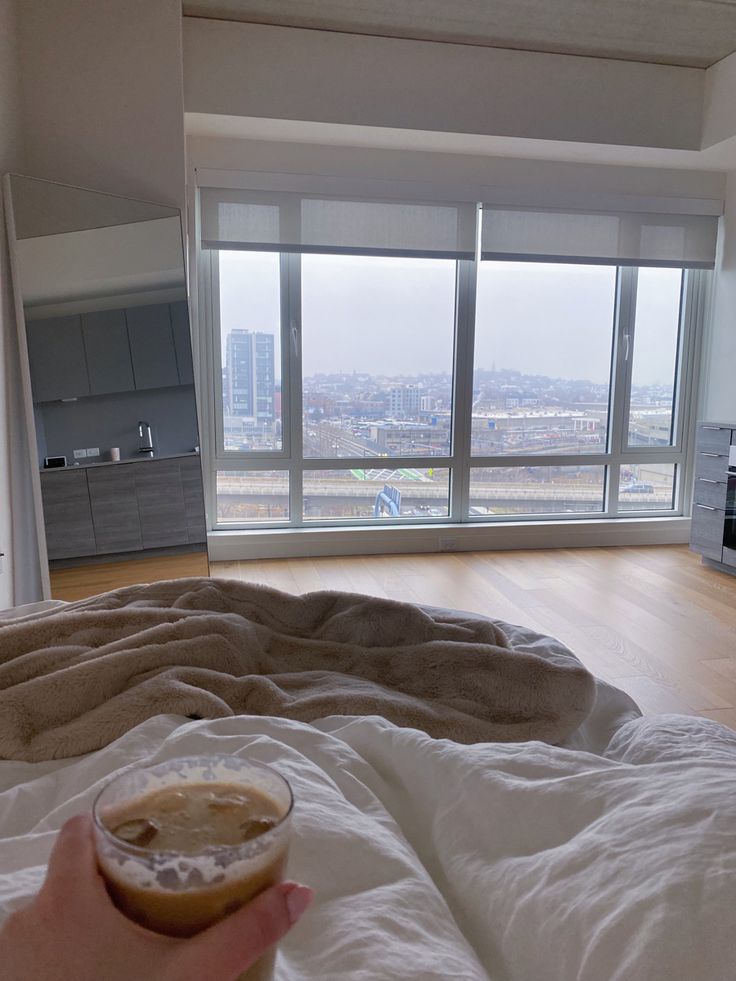 a person holding a drink in their hand while laying on a bed with a city view