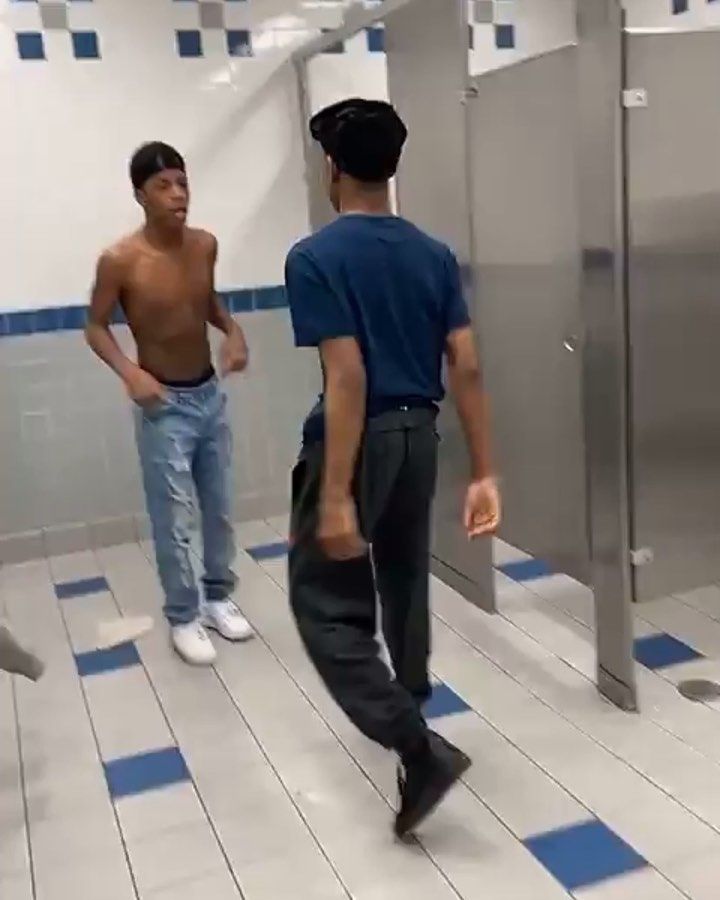 two men standing in a bathroom with blue and white tiles on the floor, one is shirtless