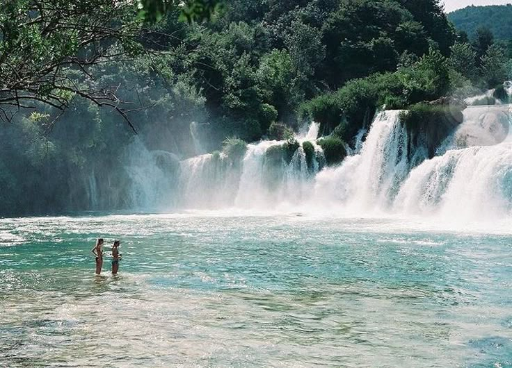 two people are standing in the water near a large waterfall with trees and bushes behind them