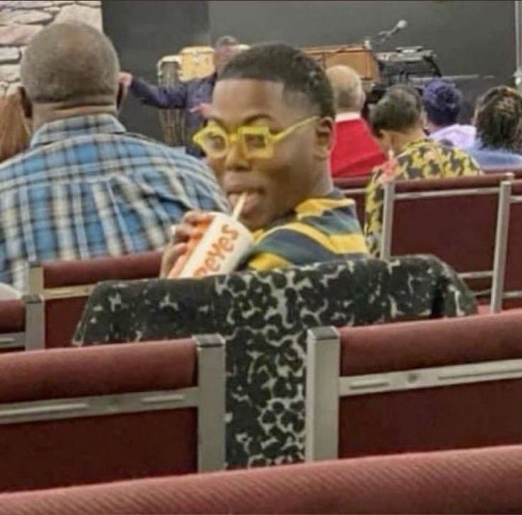 a man sitting in a chair eating a hot dog and wearing yellow glasses with other people behind him