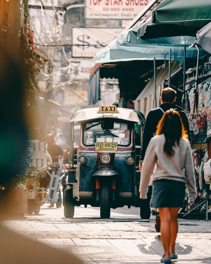 two people walking down the street in front of an old car and taxi on a busy city street