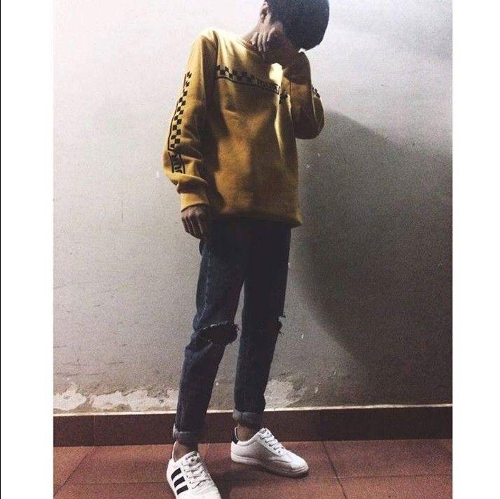 a man standing in front of a wall wearing a yellow sweater and black jeans with white sneakers