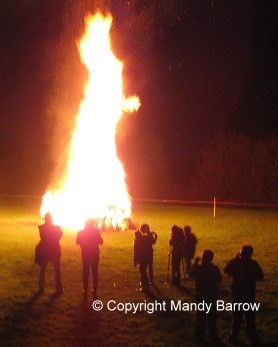 people standing around a large fire in the middle of a field at night with bright lights