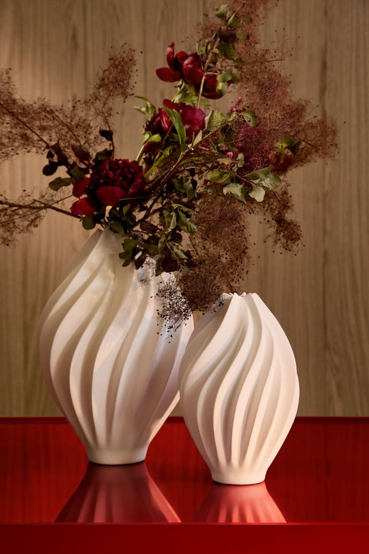 two white vases with flowers in them sitting on a red table next to a wooden wall