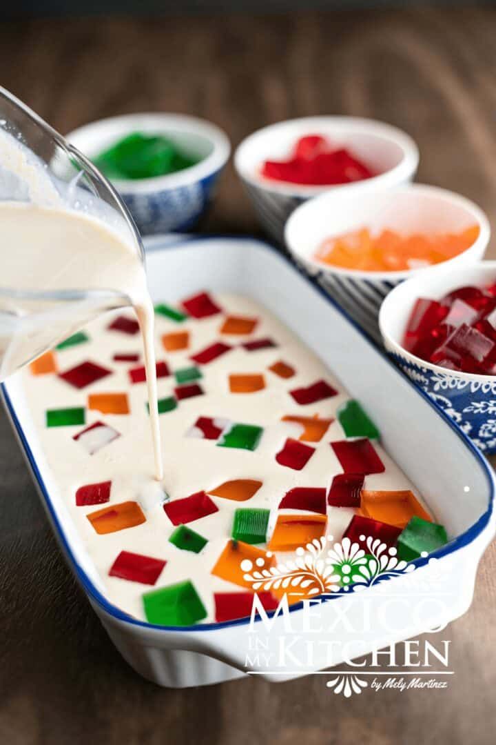 a person pouring white liquid into a dish filled with colorful jello