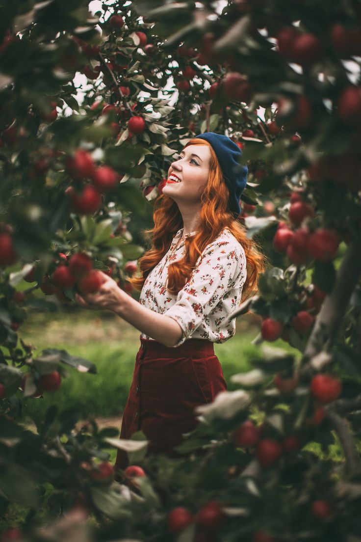 a woman standing in an apple orchard holding onto the fruit on the tree and looking up