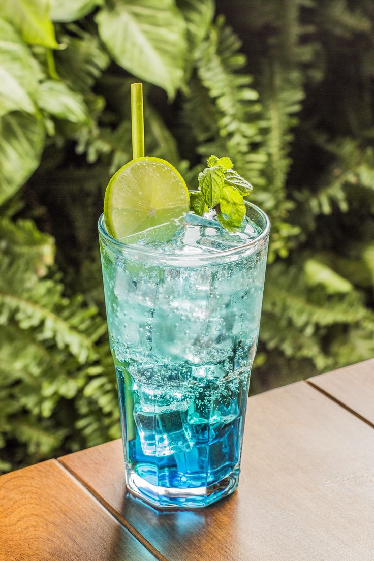 a glass filled with blue liquid and a lime wedge on top of the glass, sitting on a wooden table in front of green foliage