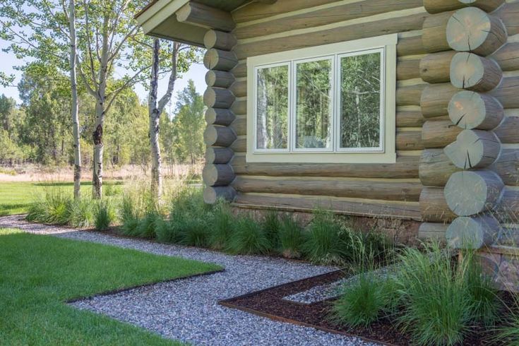 a log cabin with grass and flowers in the foreground, next to a gravel path