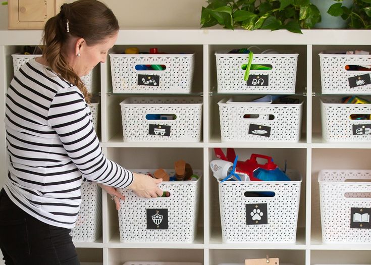 a woman standing in front of a white shelf filled with bins and boxes full of toys