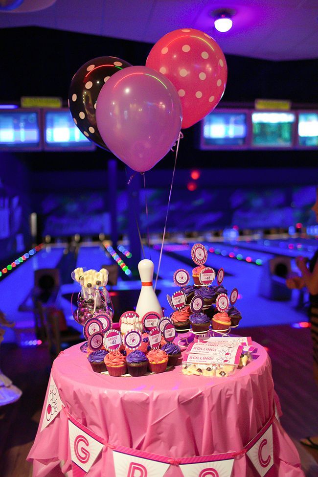 a pink table topped with lots of cupcakes and balloons next to a bowling alley