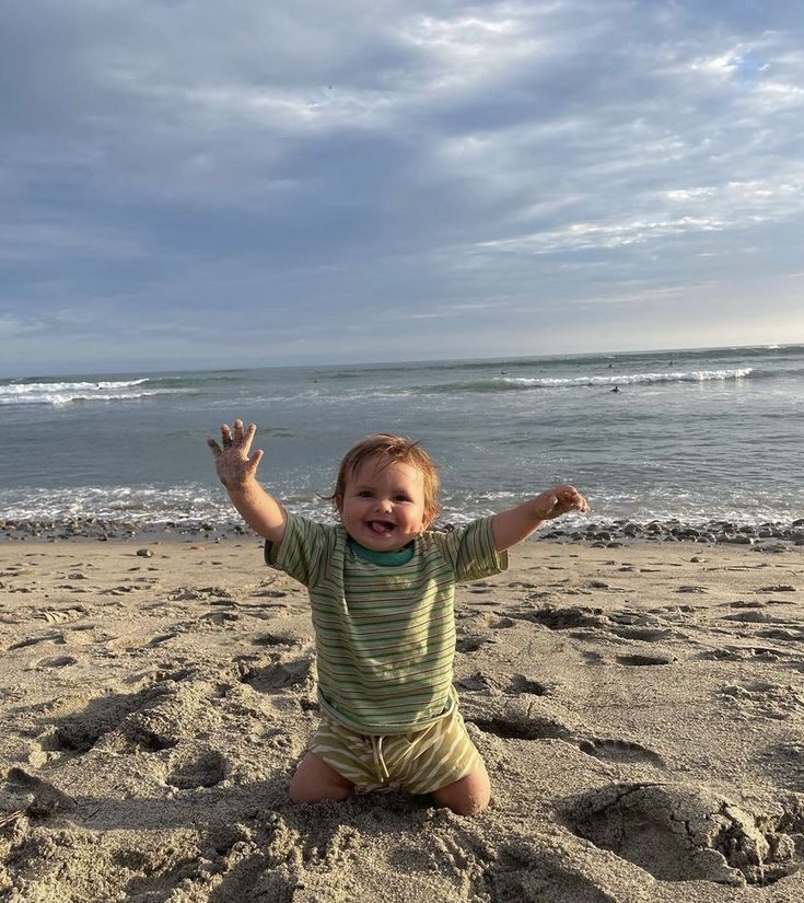 a baby sitting on the beach with his arms in the air and one hand up