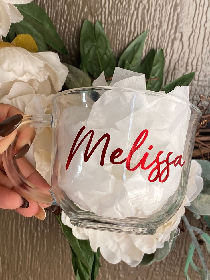 a person holding a glass with the word melsia on it and flowers in the background