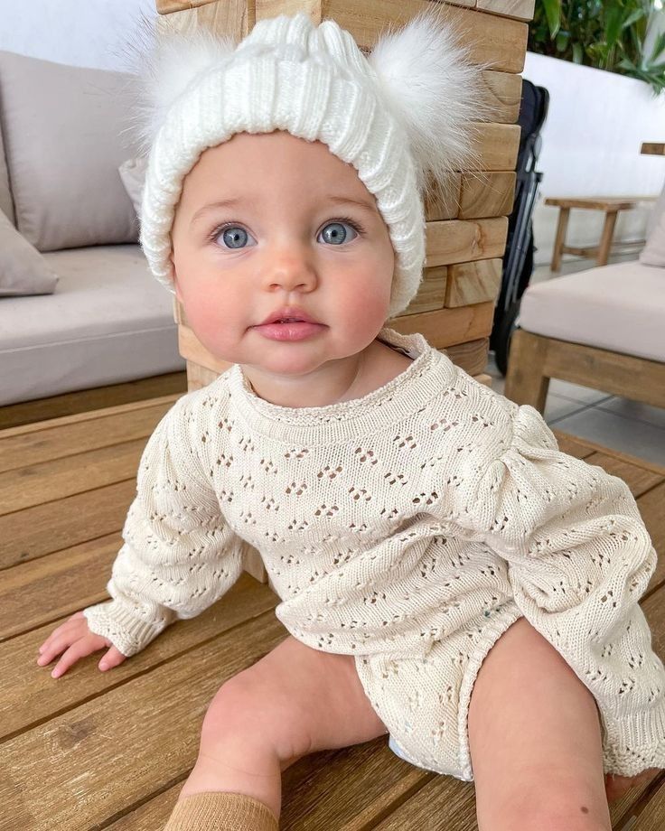 a baby sitting on a wooden bench wearing a white knitted outfit with fuzzy pom - poms