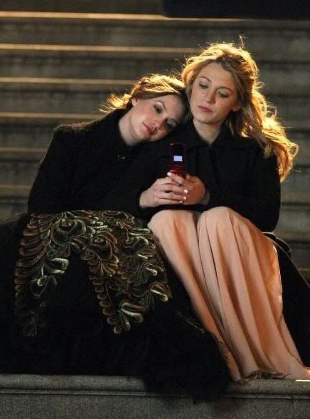 two women are sitting on the steps looking at their cell phones while one holds her arm around the other's shoulder