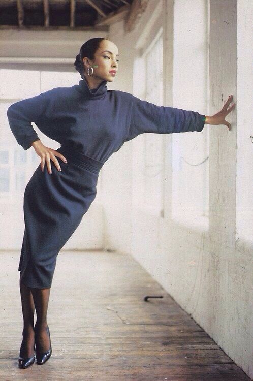 a woman leaning against a wall with her hand on the wall, wearing a blue dress and black heels