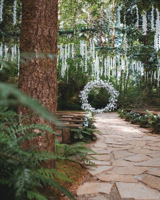 a stone path leading to a wooden bench in the middle of a forest with white flowers hanging from it