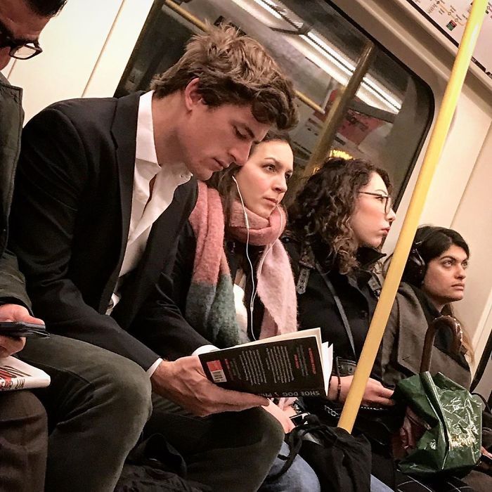 several people sitting on a subway train and one is reading a book while the other looks at his phone