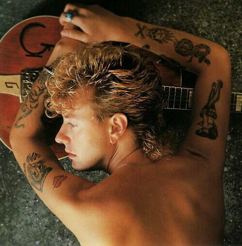 a shirtless young man holding a guitar and looking at the ground with tattoos on his arms