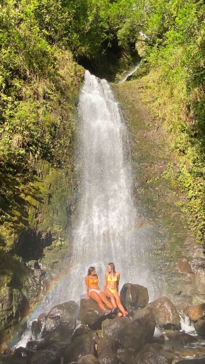 two women are sitting on rocks in front of a waterfall with a rainbow behind them