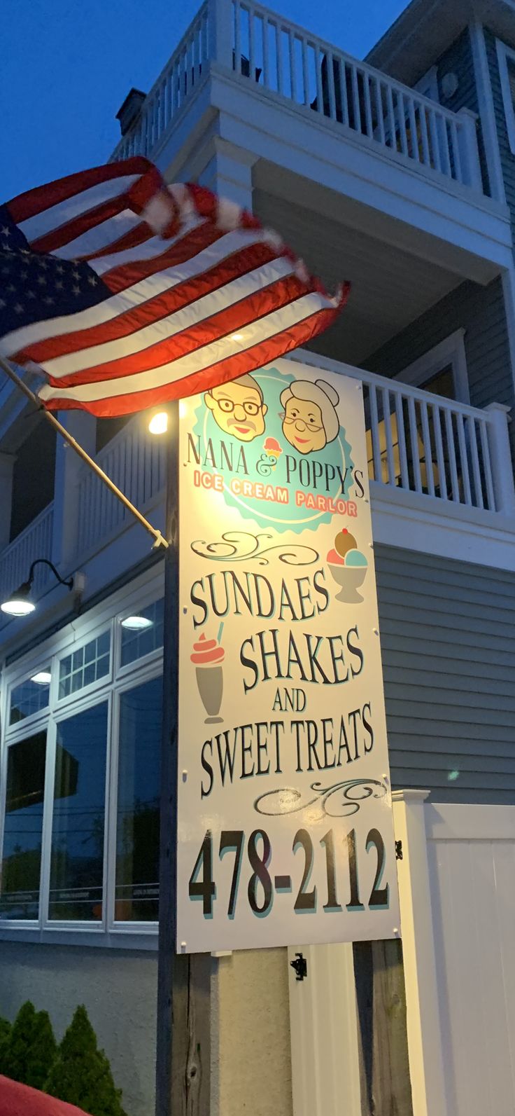 a sign with an american flag on top of it in front of a building that says sundaes shakes and sweet treats