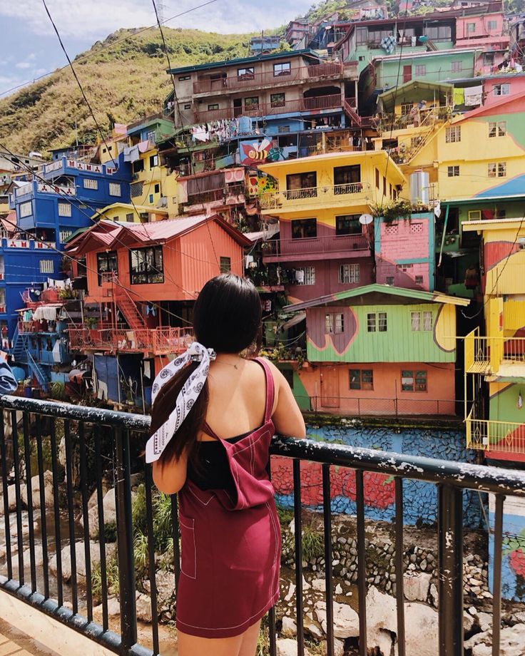 a woman standing on top of a balcony next to a metal railing with colorful houses in the background