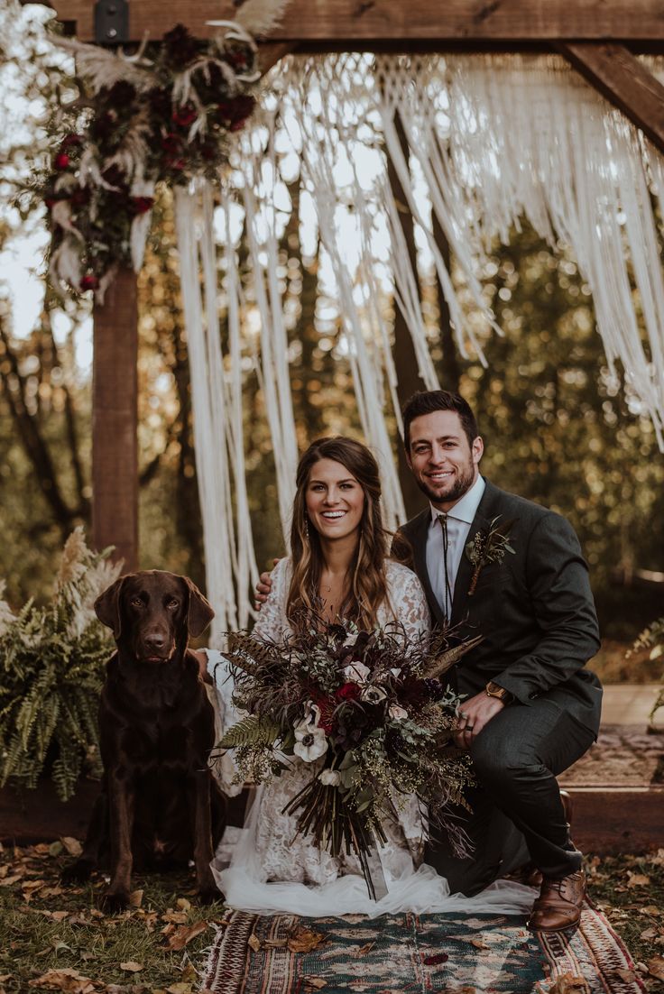 a bride and groom pose with their dog in front of an outdoor wedding ceremony arch