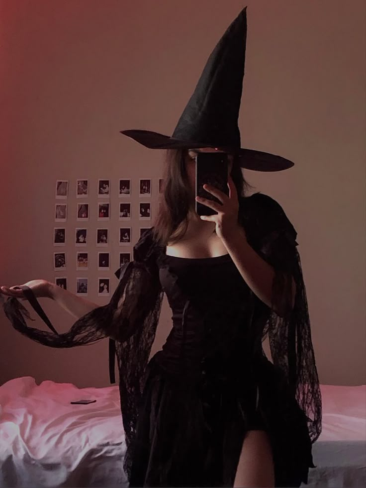 a woman in a witch costume taking a selfie with her cell phone while sitting on a bed