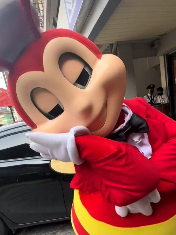 a person dressed as mickey mouse standing in front of a building with cars behind him