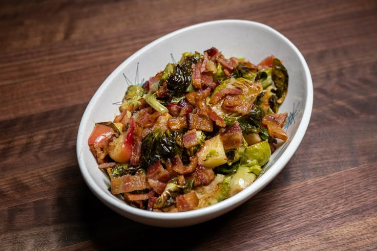 a white bowl filled with bacon and brussel sprouts on top of a wooden table