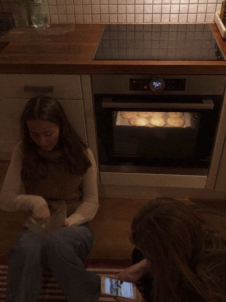 two women sitting on the floor in front of an oven looking at their cell phones