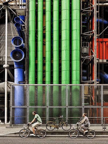 two bicyclists ride past an industrial building with pipes