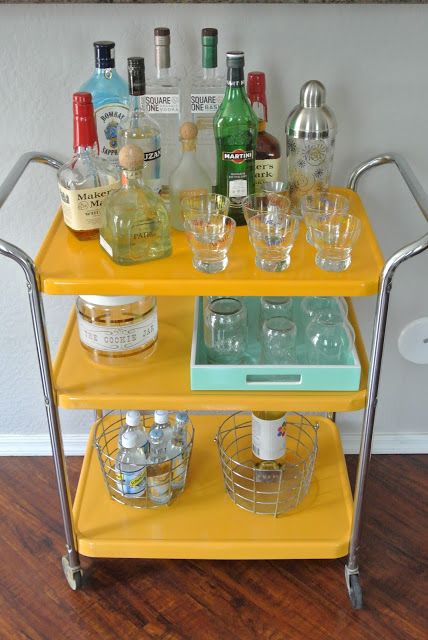 a yellow bar cart with liquor bottles and glasses on the bottom shelf next to it