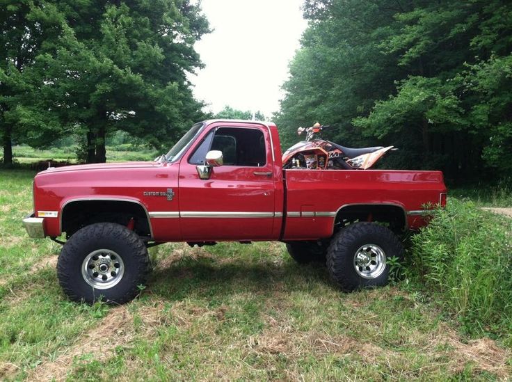 a red pickup truck parked on top of a grass covered field next to trees and bushes