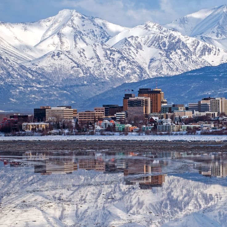 a city with mountains in the background and snow on the ground, reflecting in water