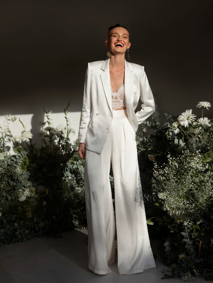 a woman standing in front of flowers and greenery wearing a white suit with wide legs