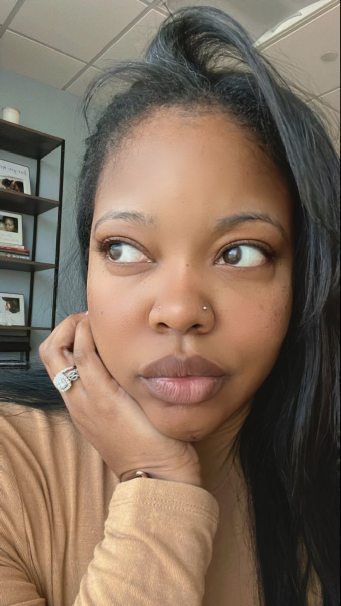 a woman with long black hair wearing a tan shirt and looking at the camera while holding her hand on her chin