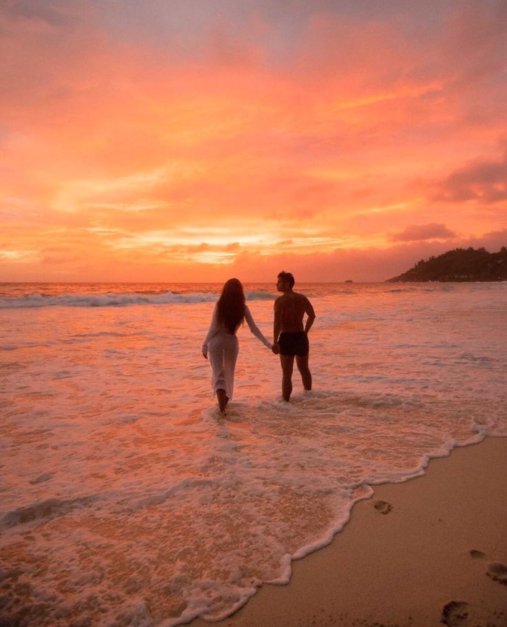 two people walking into the ocean at sunset with their hands in each other's pockets