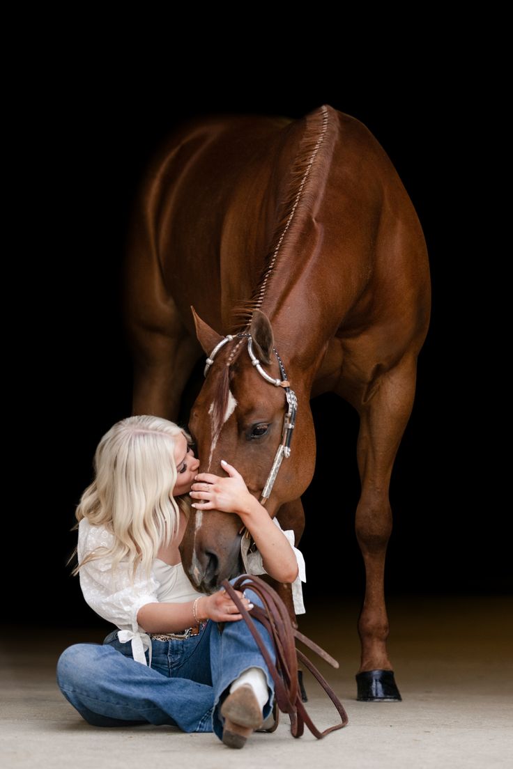 a woman kneeling down next to a brown horse with its head on it's hind legs