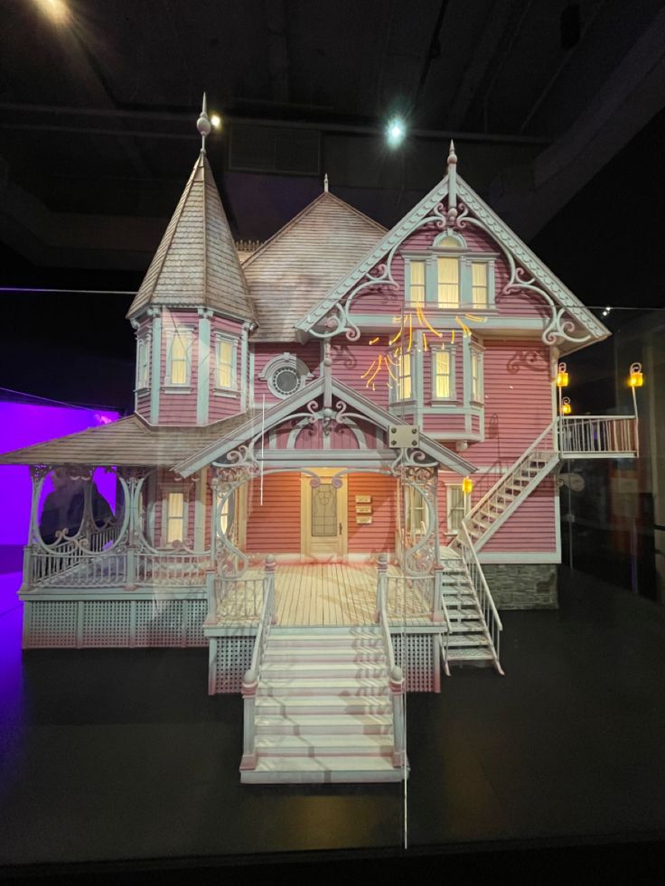 a model of a pink house on display in a museum setting with stairs leading up to the second floor