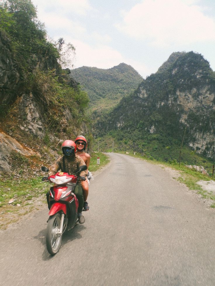 two people riding on the back of a red motorcycle down a road in front of mountains