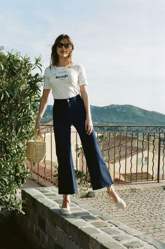 a woman in white shirt and black pants standing on brick wall next to bush with trees