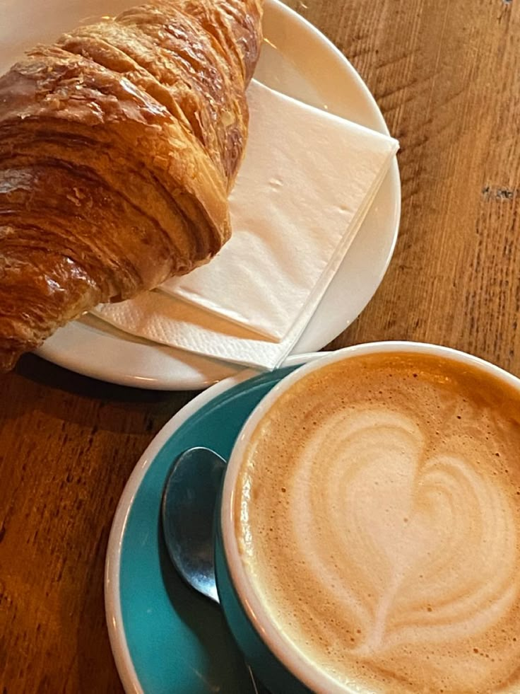 there is a croissant on the plate next to a cup of cappuccino