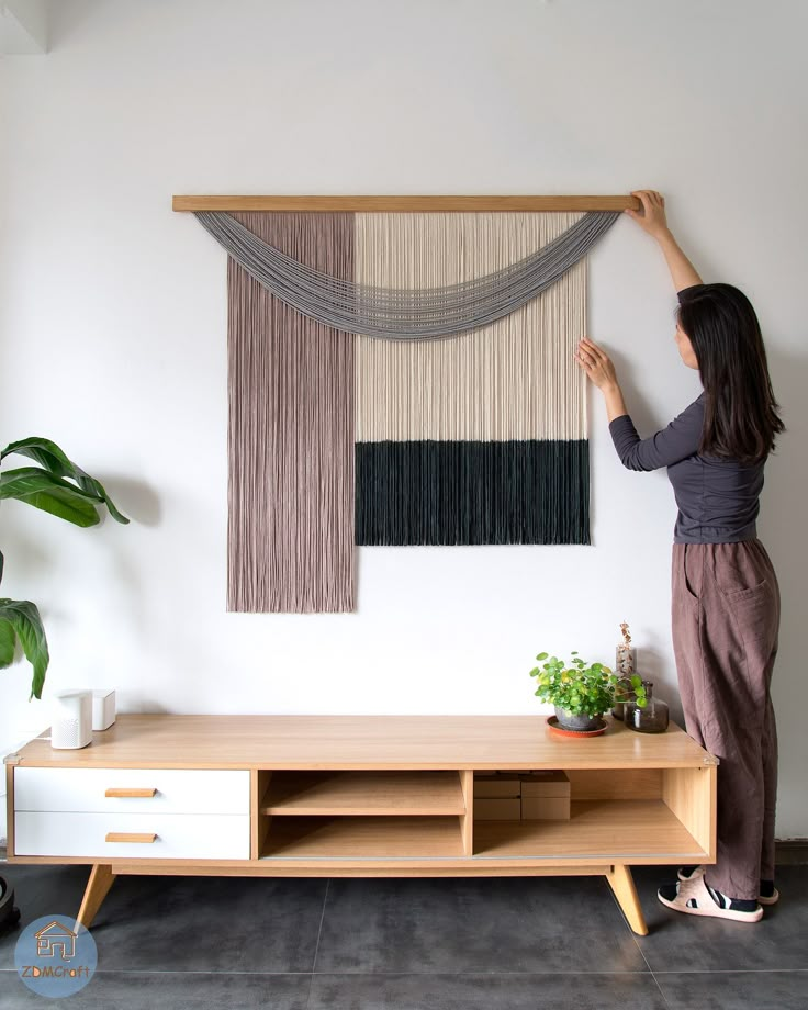 a woman is hanging curtains on the wall above a tv stand with a plant in front of it