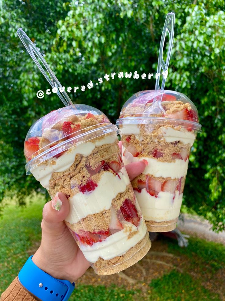 two desserts in plastic containers with strawberries and cream on top are being held by someone's hand