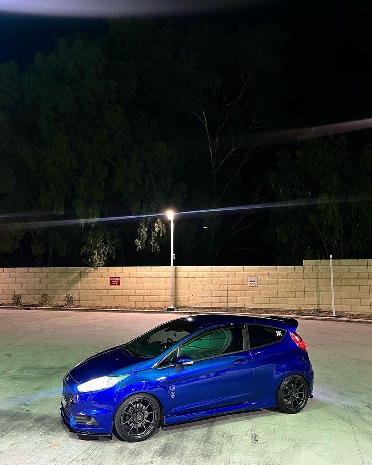 a blue car is parked in an empty parking lot at night with its lights on