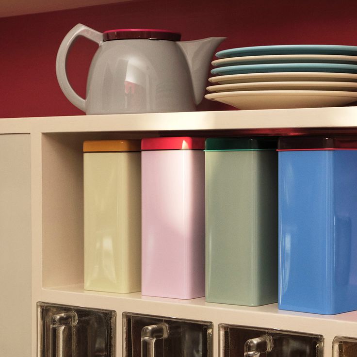 several different colored containers on a shelf with plates and teapots in the background