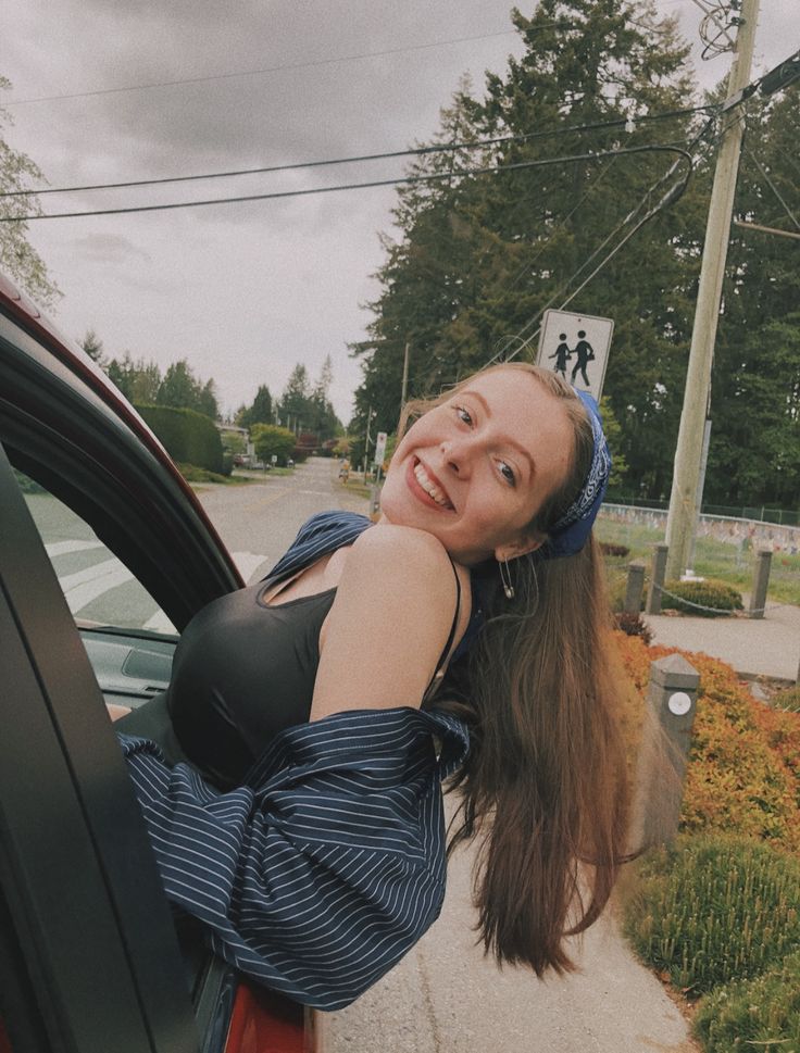 a young woman leaning out the window of a car on a street with trees in the background