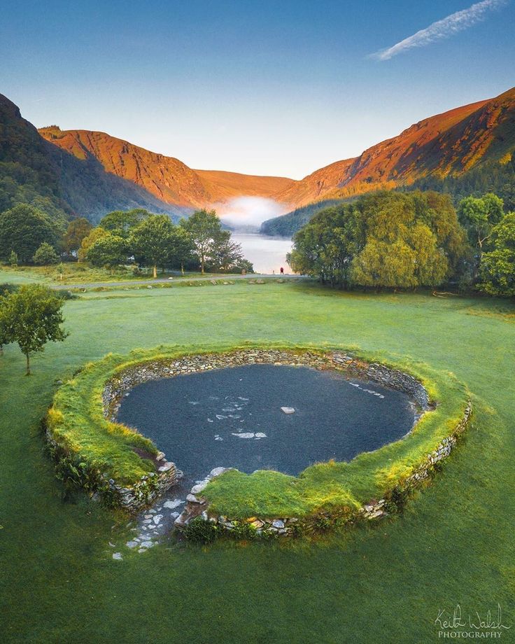 an aerial view of a pond in the middle of a green field with mountains in the background