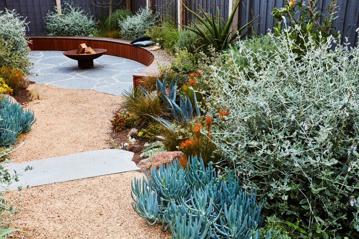 a circular bench sitting in the middle of a garden next to a gravel path and shrubbery