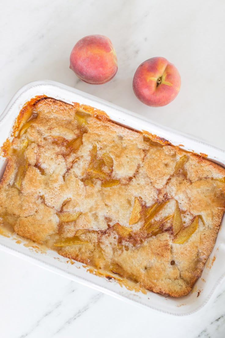 peach cobbler in a white baking dish with two peaches on the table behind it